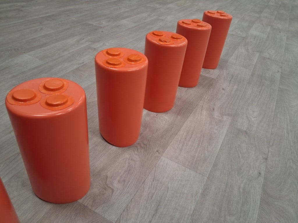 Five large orange Lego-like cylinders with four studs on top, lined up diagonally on a wooden floor.